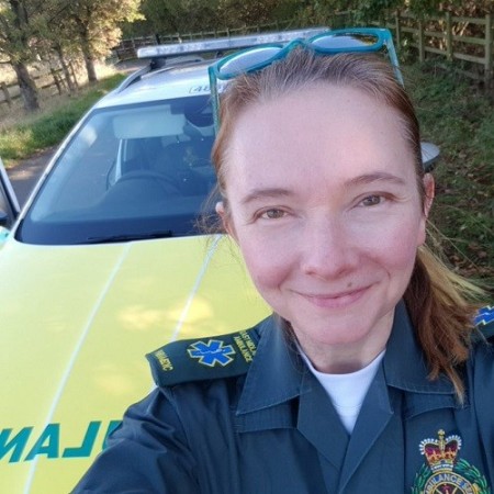 Tamsin Ryan in ambulance uniform stood smiling in front of a solo response car driven by paramedics. She is 