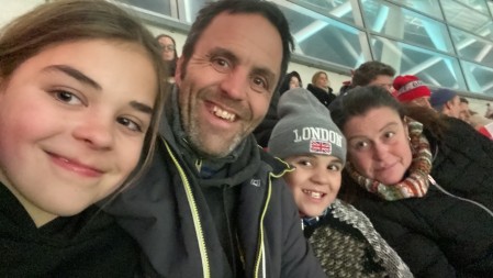 Grace and Maisie Tooley sat with their parents Adam and Charlie Tooley. They are at a sports event in the audience. They all smile for a selfie picture.