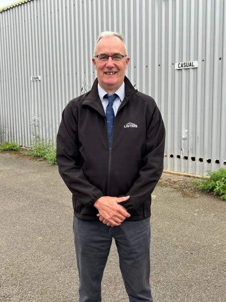 Nick Tooby wearing a black jacket stands confidently in front of his workplace, showcasing a professional demeanor. He has short grey hair and is wearing glasses.