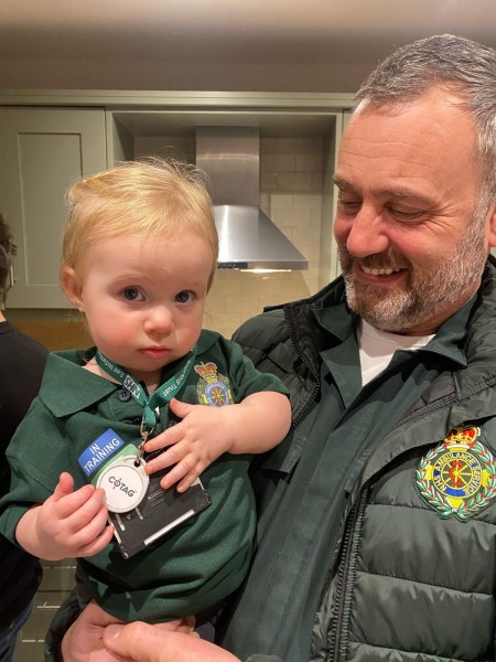 Dave Williams pictured as he is today, in the modern green ambulance uniform. He has a beaming smile while holding his granddaughter, who is wearing a mini-green ambulance t-shirt with a message on it which says "in training". 